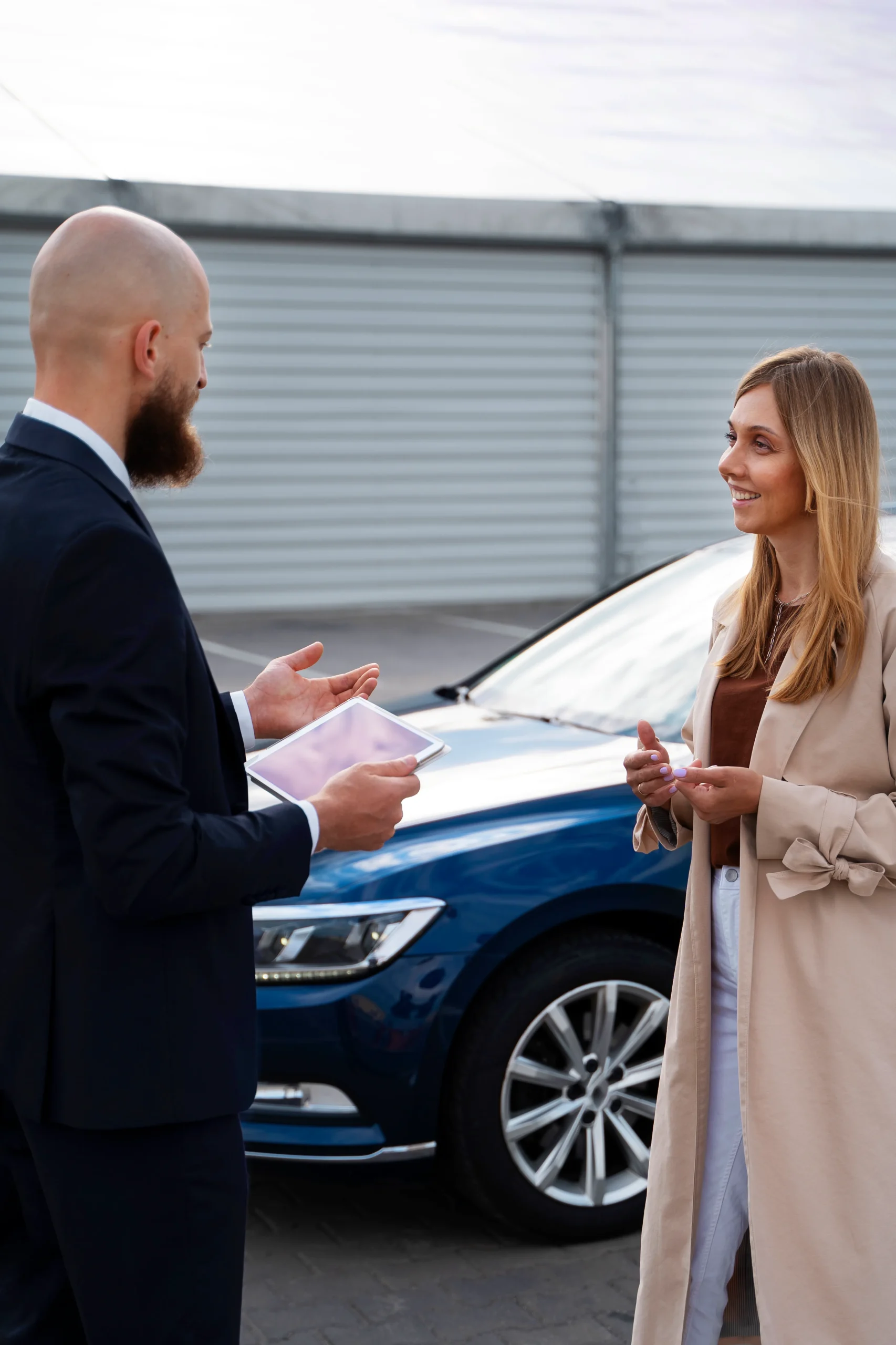 Smiling woman talking to a man with a tablet in front of a blue car in a parking lot.